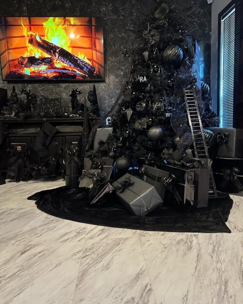 Black Christmas tree with dark ornaments and gifts beside a digital fireplace.