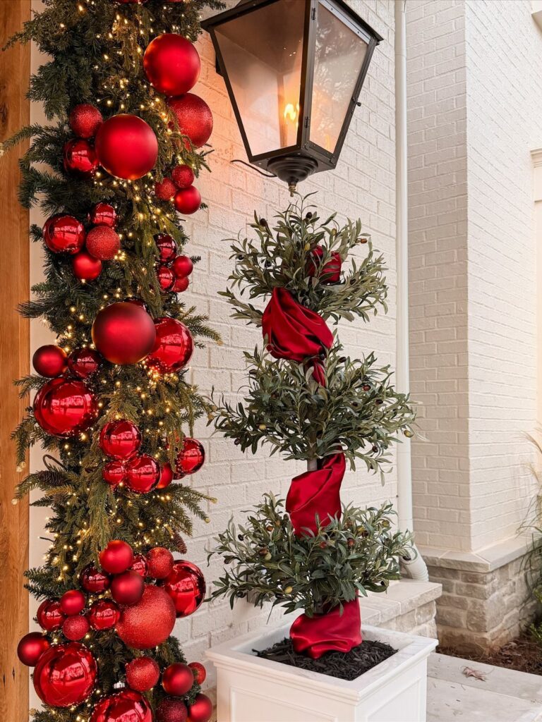 Christmas porch decor featuring a red ornament garland and ribbon-wrapped potted topiary.