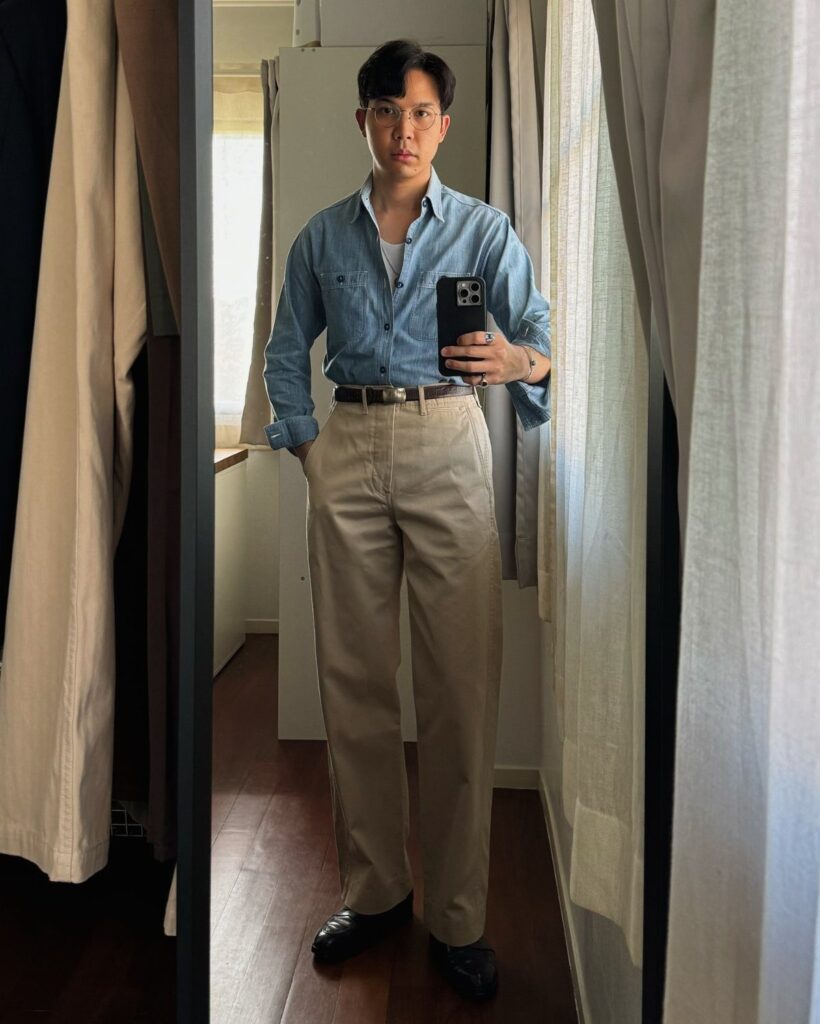Man wearing a blue chambray shirt and beige trousers taking a mirror selfie.