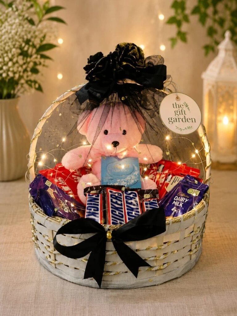 Gift basket with pink teddy bear, chocolates, and fairy lights under a clear dome.