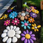 Painted stones arranged as colorful flowers on dark garden soil near green grass