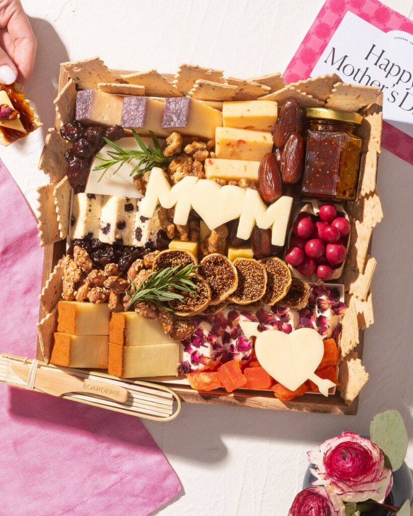 Wooden cheese board with assorted cheeses, fruits, and nuts featuring MOM cheese cutouts.