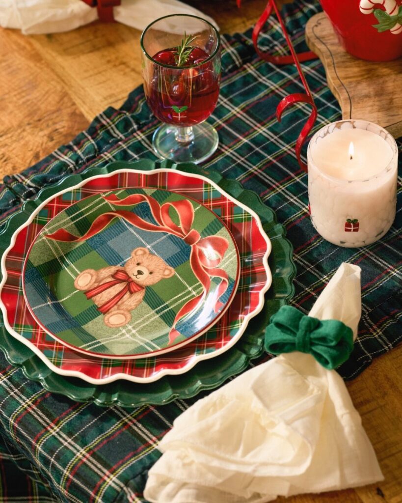 Plaid Christmas table setting with teddy bear plates, red drink, and lit candle.